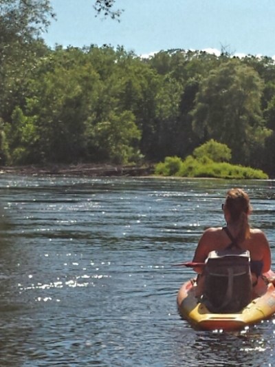 Two people kayaking on a calm river with a forested shoreline under a clear blue sky.
