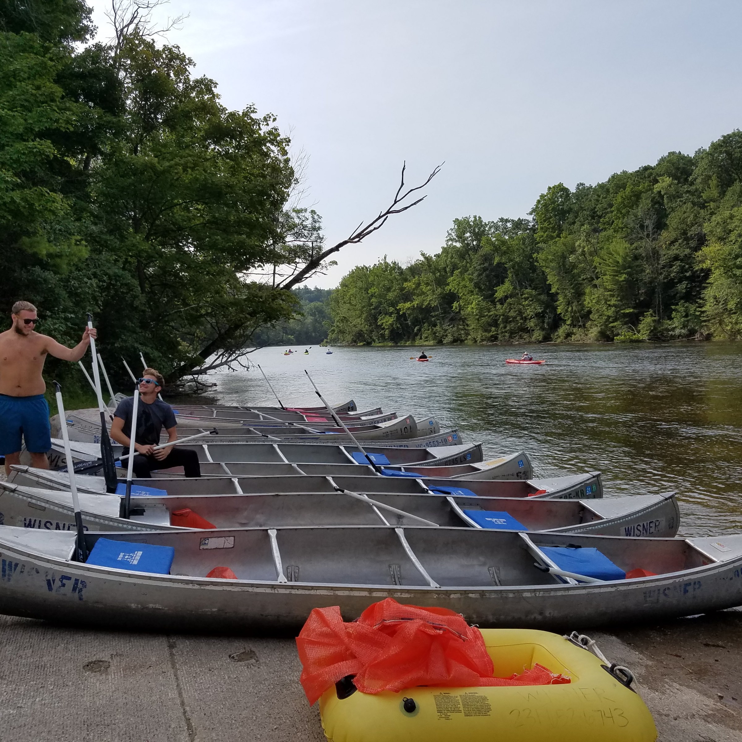Two people with paddles by canoes on a riverbank with trees in the background.
