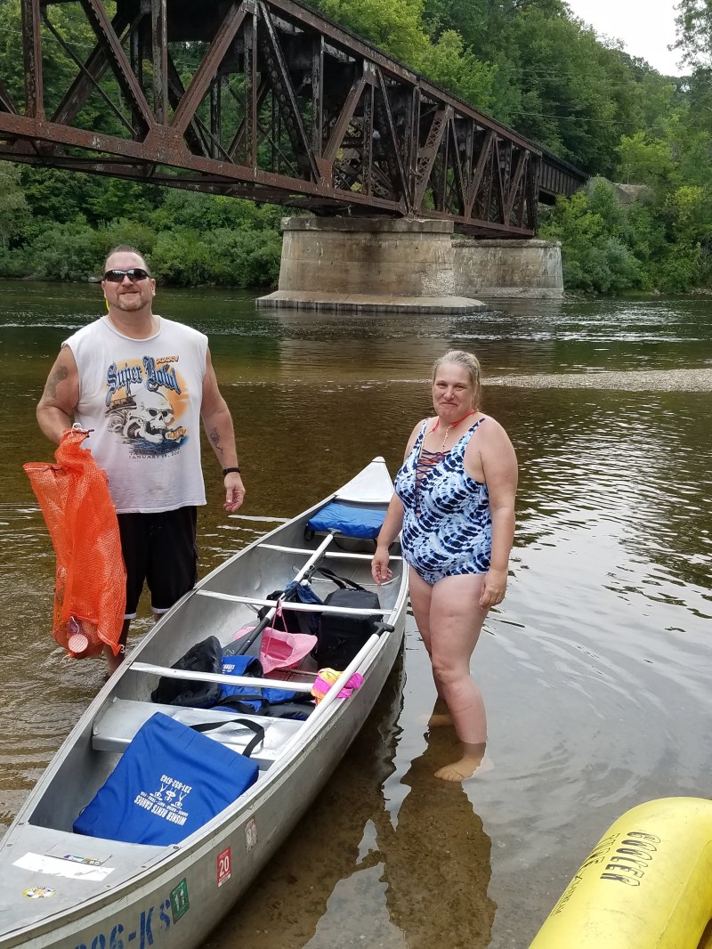 Two people stand by a canoe in a river, with a bridge and trees in the background.