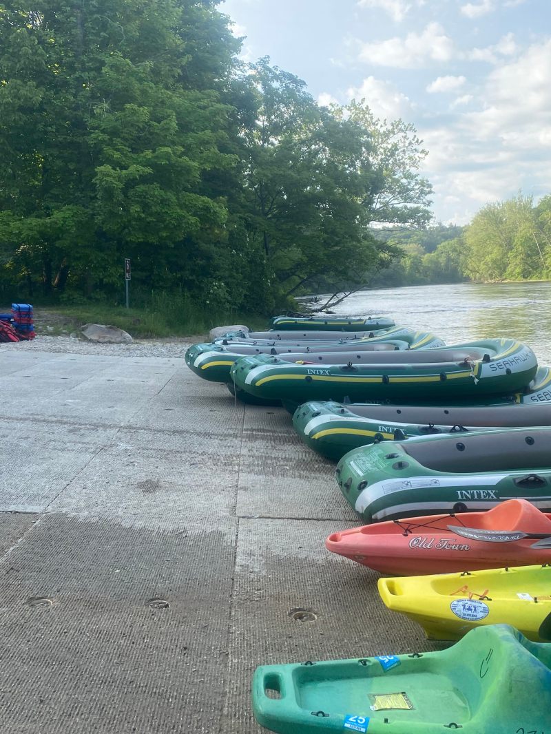 Inflatable boats and kayaks lined up by a river with trees in the background.