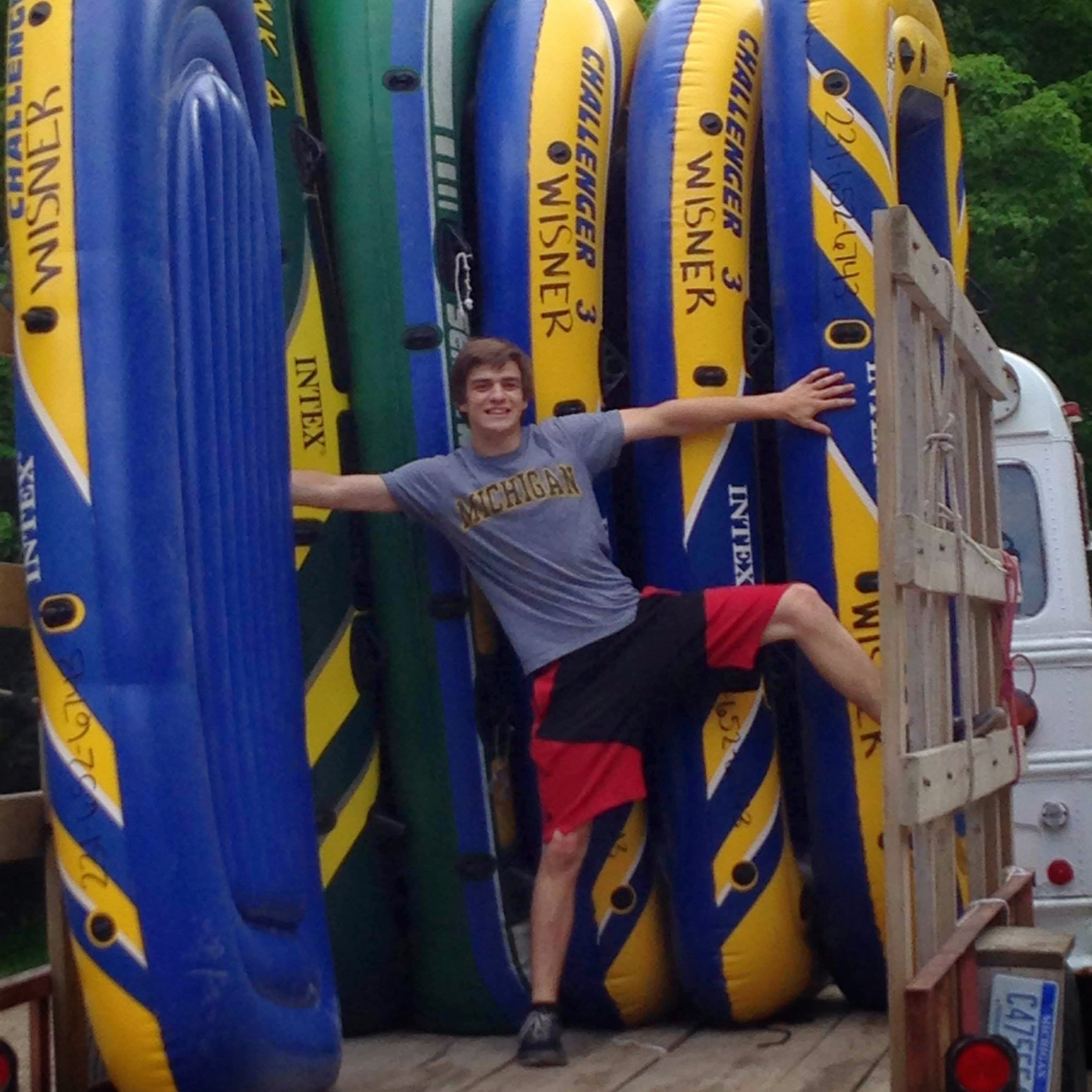 Person posing with arms outstretched among several stacked inflatable boats on a trailer.