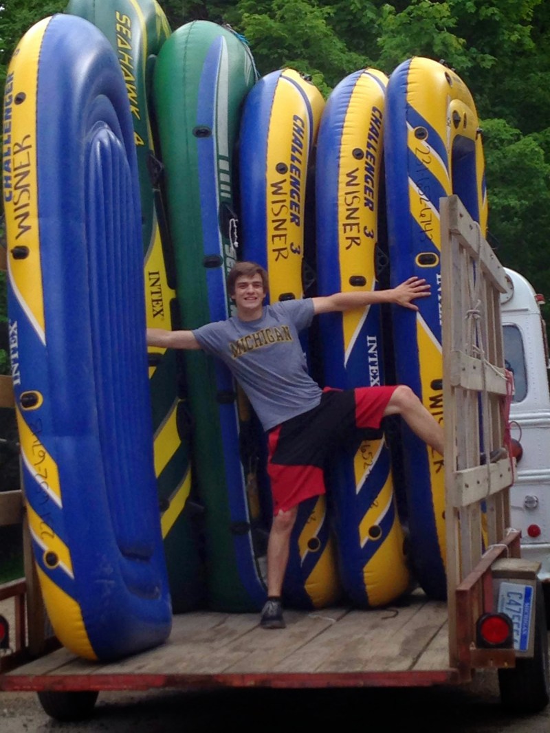 Person posing with arms outstretched among several stacked inflatable boats on a trailer.