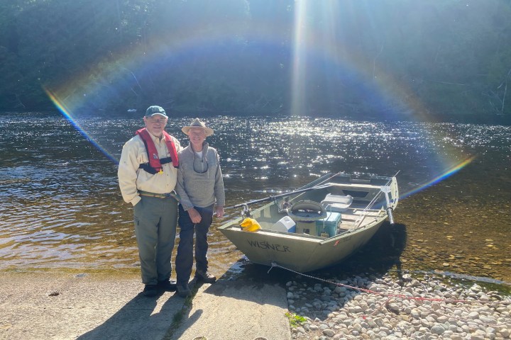 Two people stand by a boat on a sunny riverbank with a rainbow in the sunlight.