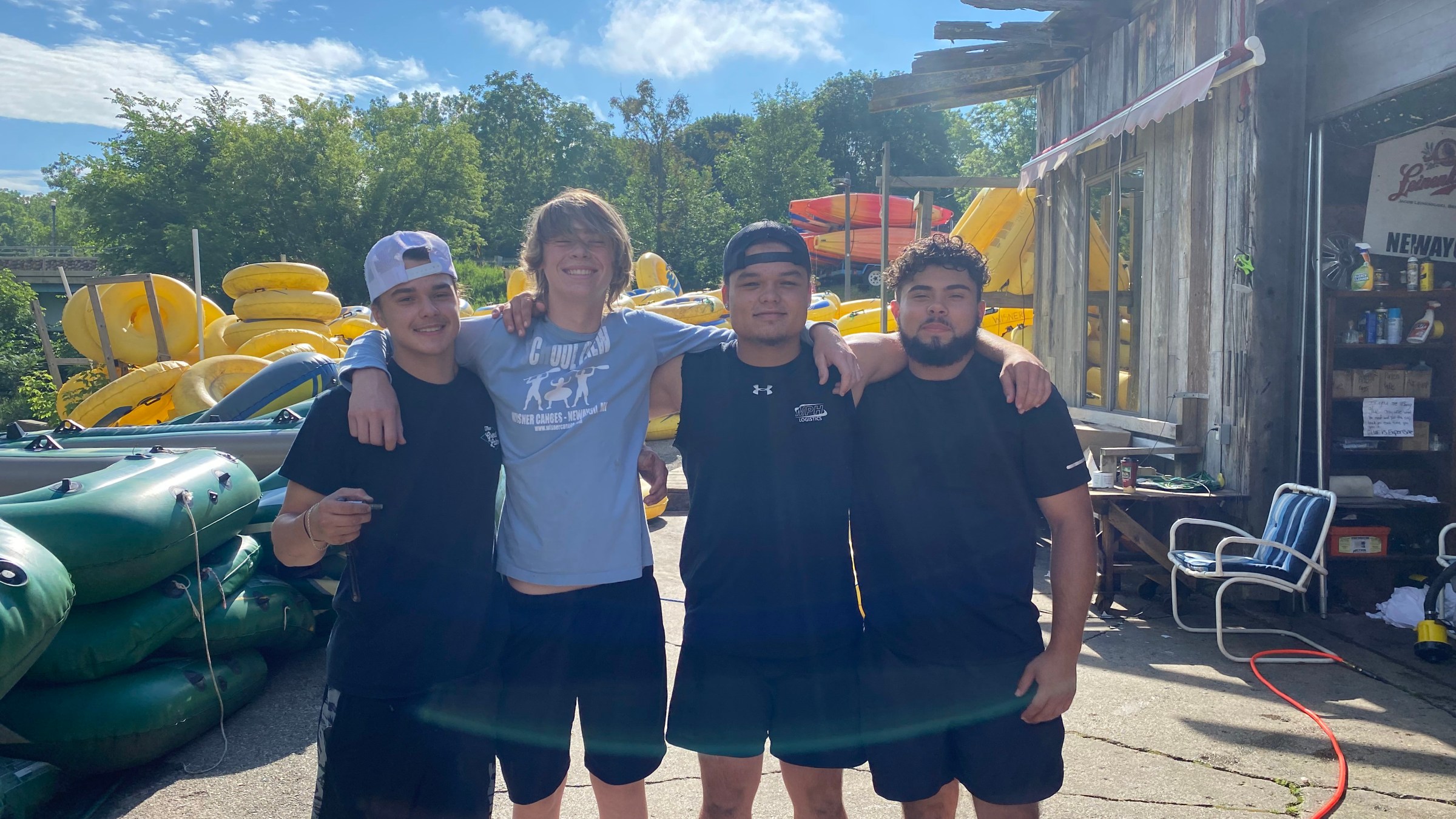 Four young men standing arm-in-arm outside near stacks of inflatable tubes and greenery.