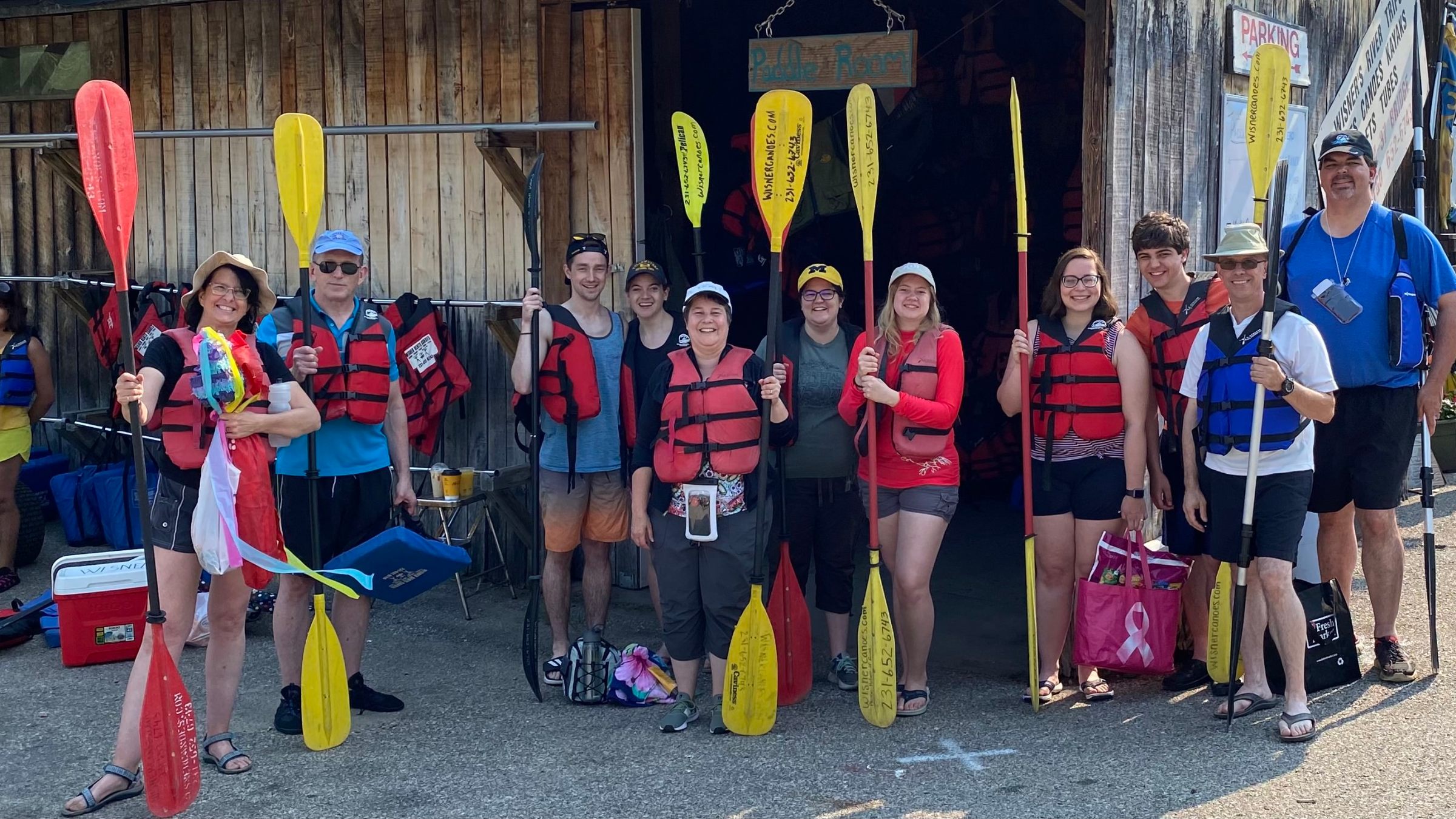 Group of people wearing life jackets holding paddles in front of a wooden shack.