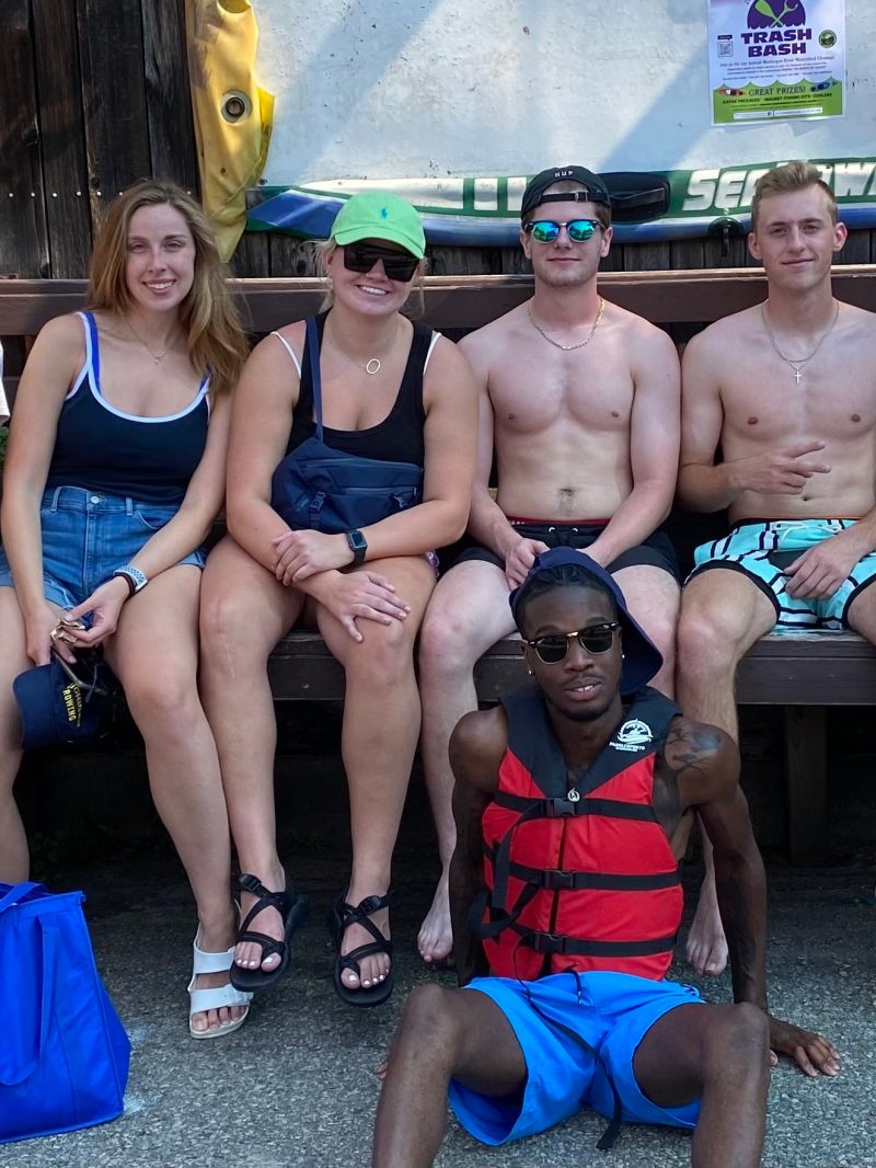 Group of young people in summer attire, sitting and standing, some wearing swimsuits, in outdoor setting.