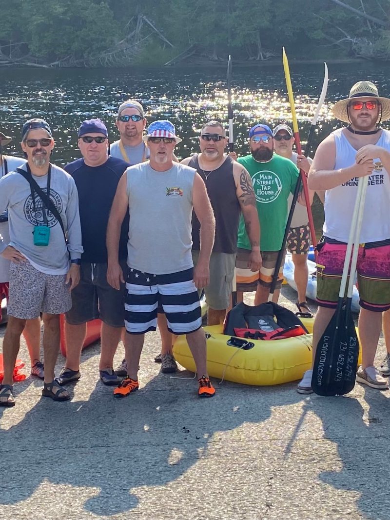 Group of men in casual summer attire with kayaks by a river.