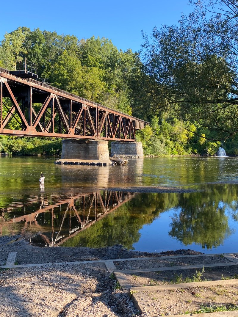 Rustic bridge over a calm river with trees and a bright blue sky in the background.