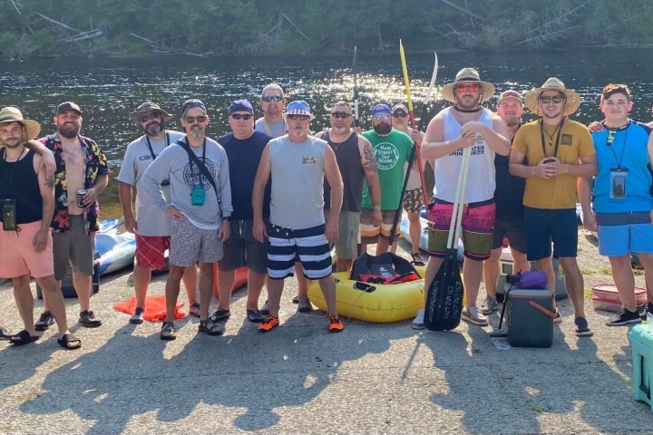 Group of men in summer clothing posing with kayaks by a river.
