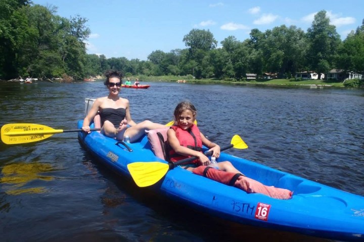 Two people kayaking on a river, surrounded by greenery on a sunny day.