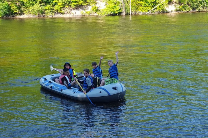 Four kids on an inflatable raft in a river, wearing life vests and waving.